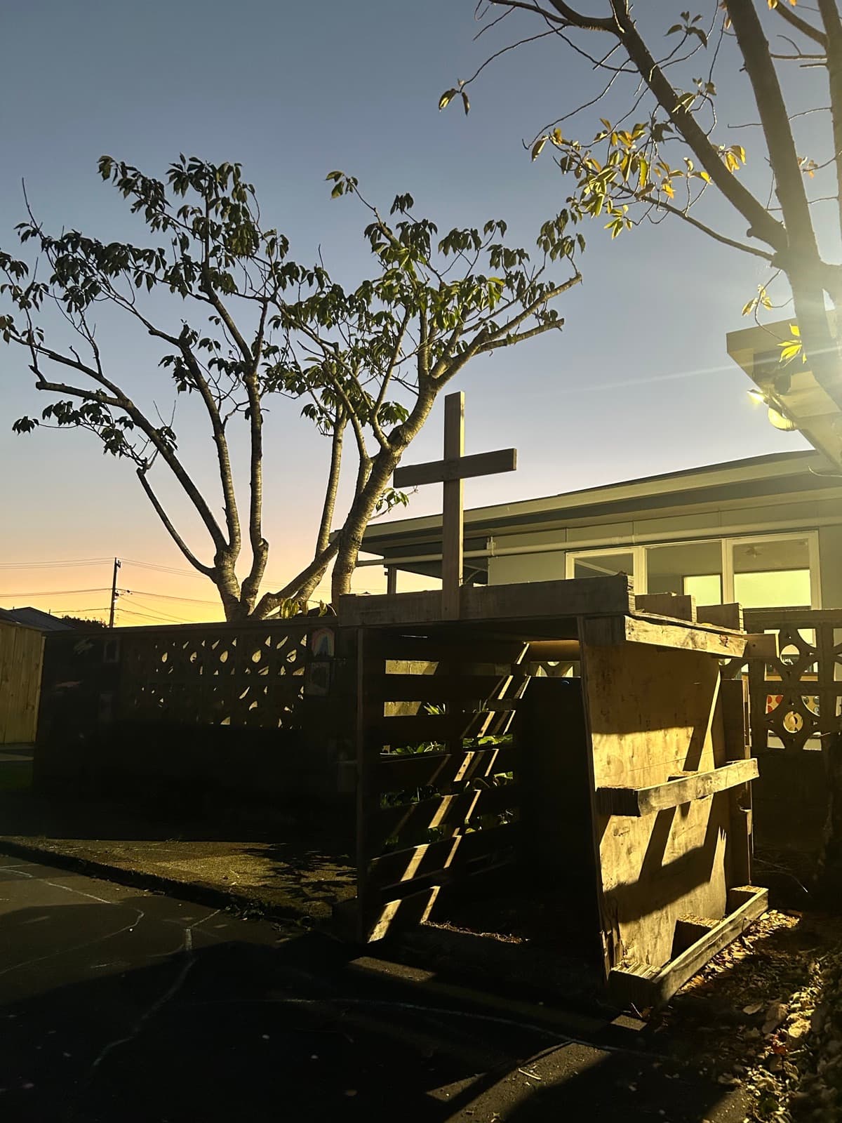 The wooden cross outside St Matt's at dawn, with the church and a tree lit by early morning sun